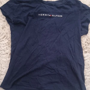Mörkblå t-shirt från Tommy Hilfiger - Snygg mörkblå t-shirt från Tommy Hilfiger med klassisk logga broderad på bröstet. T-shirten har rund halsringning och korta ärmar. Tillverkad i mjuk bomull som är skön att bära hela dagen. Den har ett otroligt litet hål ungefär som en myra nere vid sömmen men man ser det inte. Den kom tyvärr till mig så. Trycker in bra skick pga hålet annars är det mycket bra skick🩷👍