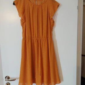 Orange kortklänning med knyt i midjan stl M - Superfin orange kortklänning med diskret prickigt mönster och volangärmar. Klänningen har rund halsringning, knytband i midjan och lätt utställd kjol. Perfekt för dig som gillar färg och vill ha en luftig och bekväm stil. Lapp bortklippt innan jag hann använda den.