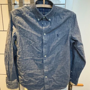 Blå skjorta från Ralph Lauren - Säljer en klassisk blå skjorta från Ralph Lauren med button-down krage och broderad logga på bröstet. Skjortan har långa ärmar och knäppning framtill. Perfekt för en stilren look. Pris kan diskuteras.