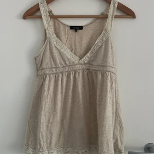 Beige linnetopp med spets från Cubus - Säljer en söt beige linnetopp från Cubus med v-ringning och spetsdetaljer längs axelbanden och nederkanten. Toppen är ärmlös och har en avslappnad passform, perfekt för varma dagar. Tillverkad i mjuk bomull för en skön känsla.