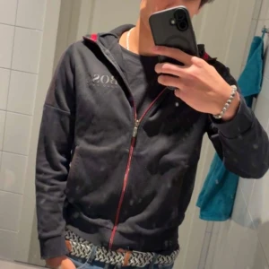 Svart zip-hoodie från Hugo Boss - Svart hoodie från Hugo Boss med röd dragkedja och logga på bröstet. Den har Snygga röda detaljer på insidan av huvan och längs dragkedjan. Perfekt för en snygg och stilren look.