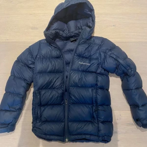 Mörkblå Peak Performance dunjacka - Säljer en mörkblå dunjacka från Peak Performance med huva och dragkedja. Jackan är quiltad och har en clean look med diskret logga på bröstet. Perfekt för kalla dagar och riktigt skön att bära. Passar dig som gillar stilren och sportig vibe. Skick 9/10 som ny