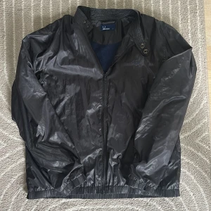 Svart vindjacka från Fred Perry L - Svart vindjacka från Fred Perry i storlek L. Jackan har en glansig finish, ribbad krage med tryckknappar och dragkedja framtill. Innerfodret är i mesh och jackan har elastiska muddar vid ärmslut och midja. Klassisk Fred Perry-logga broderad på bröstet. Nypris 2500, säljer för 500kr