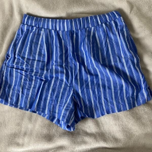 Blå randiga shorts från Gina Tricot - Snygga blå shorts från Gina Tricot i storlek S med vita vertikala ränder. De har elastisk midja och två fejk bakfickor. Materialet är mjuk bomull som känns skönt mot huden. Perfekt för varma dagar och chill stunder. Fräscha och trendiga för dig som gillar färg och mönster.