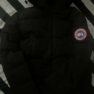Svart dunjacka från Canada Goose - Säljer en svart dunjacka från Canada Goose med klassisk patch på ärmen. Jackan är tjockt vadderad, har huva och stängs med både dragkedja och knappar. Perfekt för kalla vinterdagar och har en clean, stilren look.