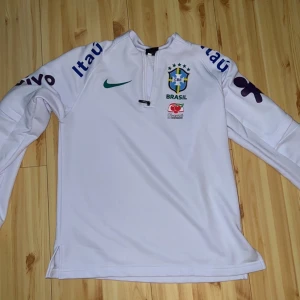 Vit Brasilien Nike fotbollströja långärmad - Vit långärmad fotbollströja från Nike med Brasilien CBF-logga på bröstet, grönt Nike-märke och sponsorer tryckta på ärmar och framsida. Tröjan har halv dragkedja och är tillverkad i ett sportigt, lätt material som andas.