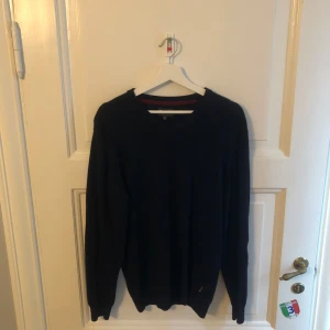 Mörkblå Barbour stickad tröja XL - Mörkblå stickad tröja från Barbour i storlek XL. Tröjan har v-ringning, långa ärmar och snygga förstärkningar på armbågarna i klassiskt rutigt tyg. Perfekt för dig som gillar stilren och tidlös design.