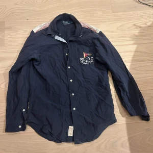 Mörkblå skjorta från Polo Ralph Lauren - Mörkblå skjorta från Polo Ralph Lauren med broderad RL-YC logga och flagga på bröstet. Skjortan har vita knappar, lång ärm och kontrastpanel i vitt och rött över axlarna. Klassisk krage och avslappnad passform. Perfekt för en cool och sportig look.
