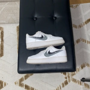 Nike Air Force 1 vita sneakers - Säljer ett par klassiska Nike Air Force 1 sneakers i vitt skinn med grå swoosh-detalj på sidorna. Skorna har rund tå, platt sula och snörning framtill. Perfekta för dig som gillar en clean och tidlös look med en twist av grått.