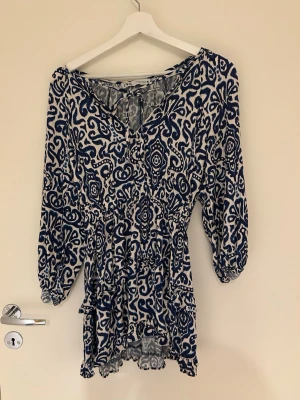 Blåvit mönstrad kortklänning Zara  - Kortklänning från Gina Tricot i blått och vitt med ett unikt grafiskt mönster. Klänningen har v-ringning, långa ärmar med resår vid ärmslut och volangdetaljer nedtill. Tillverkad i ett lätt och luftigt material, perfekt för varmare dagar.