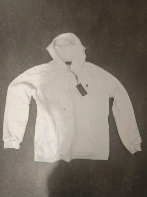 Vit hoodie Polo Ralph Lauren - Snygg vit hoodie Polo Ralph Lauren med dragkedja framtill och klassisk huva. Hoodien har fickor på magen och den ikoniska lilla loggan broderad på bröstet. Perfekt för en clean och stilren look. Materialet känns mjukt och skönt.