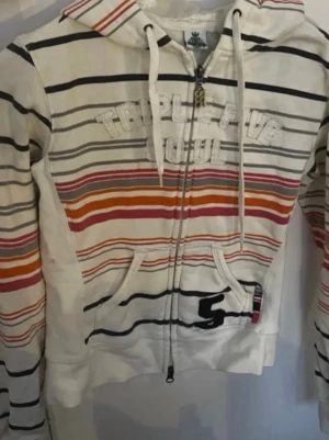 Y2K Randig hoodie från Triple Five Soul - Vit hoodie med dragkedja från Triple Five Soul. Har färgglada ränder i svart, orange, rött och rosa samt broderad logga på bröstet. Utrustad med huva och snören, samt två fickor framtill. Perfekt för en avslappnad streetwear-look.