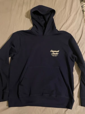 Mörkblå hoodie Originals Studio - Mörkblå hoodie från Jack n Jones med vit text på bröstet och stor print på ryggen. Klassisk känguruficka framtill och huva. Tillverkad i mjukt bomullsmaterial, perfekt för en avslappnad och stilren look.