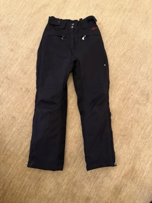 Svarta täckbyxor med dragkedjor - Svarta täckbyxor med raka ben, dragkedjor vid benslut och praktiska fickor framtill. Byxorna har bälteshällor och en liten röd detalj vid ena fickan. Perfekta för kalla dagar och utomhusaktiviteter. Passar ridning eller skidåkning. Storlek 140 / 146.