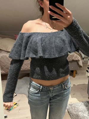 Mörkgrå off shoulder topp - Trendig mörkgrå off shoulder topp med volang runt hela axelpartiet. Toppen är croppad och har långa ärmar i mjukt, stretchigt material. Perfekt för dig som vill ha en snygg och bekväm topp med unik design.