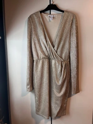 Glittrig guldklänning från NLY Trend - Säljer en supersnygg, glittrig guldklänning från NLY Trend i omlottmodell med v-ringning och långärmade ärmar. Klänningen är kort och har snygga draperingar i midjan som ger en cool look. Perfekt för dig som vill sticka ut med lite extra glitter. Använd en gång.