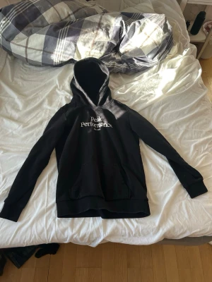 Svart hoodie från Peak Performance - Säljer en svart hoodie från Peak Performance med vit logga på bröstet. Klassisk modell med huva och känguruficka framtill. Tillverkad i mjukt material och har ribbade muddar vid ärmslut och nederkant. Perfekt för en avslappnad och stilren look.
