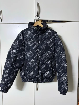 Svart pufferjacka från Ballou Couture - Snygg svart pufferjacka från Ballou Couture med vit textprint över hela jackan. Jackan har hög krage, dragkedja framtill och elastisk nederkant. Perfekt för dig som vill sticka ut med en streetwear-inspirerad look under vintern.