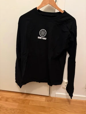 Svart långärmad Saint Pablo topp - Svart långärmad topp från Saint Pablo med vitt tryck på bröstet och längs ena ärmen. Rund halsringning och rak passform. Materialet känns som bomull och toppen har en cool grafisk detalj framtill.