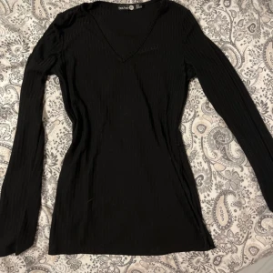 Svart ribbad långärmad topp Boohoo - Säljer en svart ribbad långärmad topp från Boohoo med v-ringning. Toppen har en slim passform och är tillverkad i ett stretchigt material som sitter snyggt på kroppen. Perfekt att styla med jeans eller kjol. Tröjan har en uppklippt del på sidorna. 