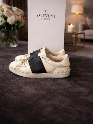 Valentino Garavani vita sneakers - Snygga vita sneakers från Valentino Garavani med bred svart läderdetalj över sidan och ovandelen. Klassisk låg modell med vita snören och subtila nitar på hälen. Perfekta för dig som gillar exklusiv streetstyle och vill sticka ut.