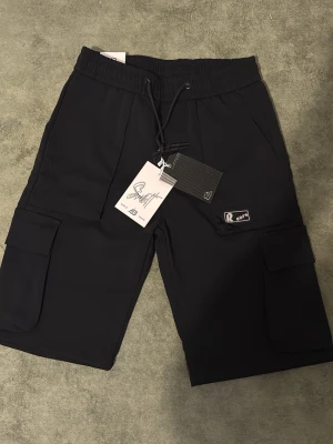 Svarta cargobyxor från Sixth June - Snygga svarta cargobyxor från Sixth June med elastisk midja och dragsko. Byxorna har flera praktiska fickor på sidorna och en patch med logga på benet. Perfekta för en streetwear-look och riktigt bekväma tack vare det mjuka materialet.