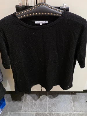 Svart glittrig topp från Flash Woman - Svart glittrig topp från Flash Woman med rund halsringning och korta ärmar. Toppens material har en lätt skimrande effekt som ger en snygg touch till din outfit. Perfekt att styla med jeans eller kjol för en trendig look.