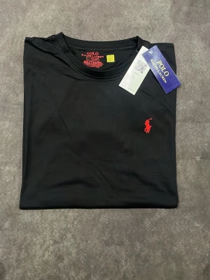 Svart t-shirt från Polo Ralph Lauren - Klassisk svart t-shirt från Polo Ralph Lauren med rund halsringning och rött broderat logotyp på bröstet. Tillverkad i mjuk bomull som känns skön mot huden. Perfekt för en stilren och enkel look.
