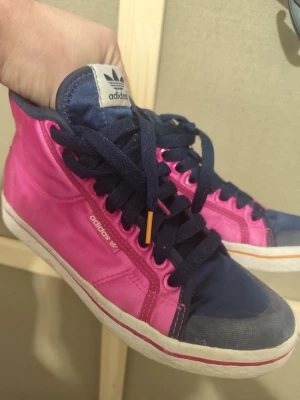 Adidas skor storlek 38,5 orange på ena sidan och rosa på andra - Färgstarka Adidas high tops med tydlig retro-känsla. Kombination av rosa, orange och mörkblå ger en riktigt nice streetwear-look som sticker ut. Använda men i helt okej skick, normala tecken på bruk. Perfekta för casual fits eller som statement-sneakers. Se bilder för skick.