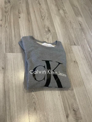 Grå sweatshirt från Calvin Klein - Snygg grå sweatshirt från Calvin Klein Jeans med stort svart CK-tryck och vit logga på bröstet. Klassisk rund halsringning och ribbade muddar vid ärmslut och nederkant. Perfekt för en avslappnad och trendig stil.