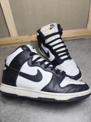 Nike Dunk High 42,5 Panda – Black White / klassisk färgsättning - Nike Dunk High i den populära Panda-färgsättningen, alltså svart och vitt i sin mest tidlösa form. En modell som funkar till allt och som alltid efterfrågas, oavsett säsong. Skorna är i bra bruksskick. Lädret har normalt slitage, främst på de svarta panelerna, men är fortfarande mjukt och helt utan sprickor eller större skador. Sulorna har bra grepp kvar och skorna känns stabila att använda direkt. Insidan visar tydliga men rimliga tecken på användning, inget som påverkar funktionen. 