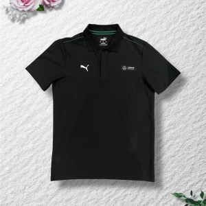 Svart pikétröja från Puma x Mercedes - Svart pikétröja från Puma i samarbete med Mercedes AMG Petronas. Tröjan har klassisk krage, korta ärmar och logotyper på bröstet. Perfekt för dig som gillar sportig och stilren design.