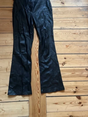 Svarta utsvängda byxor i fuskskinn - Säljer ett par svarta byxor i fuskskinn med utsvängda ben och hög midja. Snyggt glansigt material som ger en cool look och passar perfekt till streetwear eller en trendig outfit. Inga synliga fickor eller detaljer framtill.