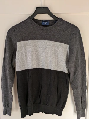 Mörkgrå stickad tröja från GANT - Snygg stickad tröja från GANT i mörkgrått, ljusgrått och svart. Tröjan har rund halsringning, långa ärmar och en bred ljusgrå panel över bröstet med diskret GANT-logga broderad. Perfekt för en stilren och avslappnad look. Mått: höjd 61 cm, axelbredd 51 cm.