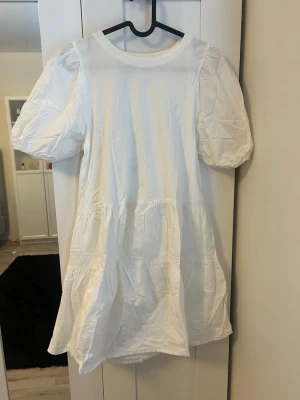 Vit kort klänning med puffärm från Zara - Säljer en vit kort klänning från Zara med puffiga kortärmar och rund hals. Klänningen har en luftig passform och volangdetaljer nedtill. Perfekt för sommarens alla tillfällen och är tillverkad i mjuk bomull.