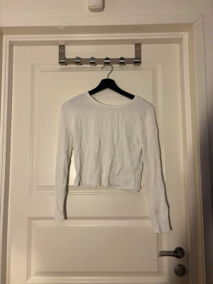 Vit ribbad croppad topp - Trendig vit långärmad topp med ribbad struktur och croppad passform. Toppen har en unik öppen detalj baktill med tre elastiska band. Perfekt för att lyfta din outfit med en cool twist. 
