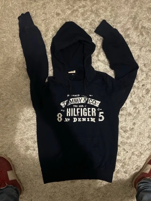 Mörkblå hilfiger hoodie - Mörkblå hilfiger denim hoodie med luva och vitt tryck samt snören. Storlek M mycket bra skick. Hör gärna av dig!