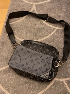 Louis Vuitton axelväska  - Säljer en snygg axelväska från Louis Vuitton i svart och grått med klassiskt monogrammönster. Väskan har justerbar axelrem i textil och detaljer i läder. Perfekt för dig som vill ha en stilren och exklusiv accessoar med plats för det viktigaste.