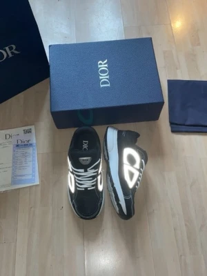 Svarta Dior sneakers med reflexdetaljer - Snygga svarta Dior sneakers med vita och reflekterande detaljer. Skorna har en chunky sula, vita skosnören och meshpaneler för extra komfort. Stora Dior-loggor på sidorna ger en exklusiv vibe. Perfekta för dig som vill sticka ut med stilrena och moderna sneakers. Boxen har några småskråmor på hörnen men annars är allting oanvänt och inga andra defekter. Hör av er vid funderingar😊
