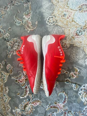 Adidas Copa röda fotbollsskor - Säljer ett par röda Adidas Copa fotbollsskor med vita detaljer och klassiska tre ränder på sidan. Skorna har en låg profil, snörning och slitstark yttersula i gummi, perfekt för inomhusspel. Materialet är syntet och textil för bra komfort och passform.