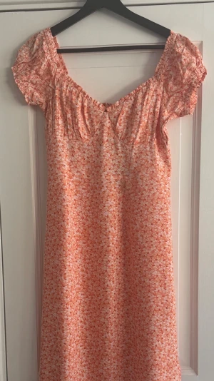 H&M klänning i stl 38 helt ny - Figurnära, vadlånga off-shoulder-klänning i vävt tyg med blommönster. Hjärtformad ringning framtill, rynkad byst och korta puffärmar. Täckt resår och volangkant runt ringning och ärmslut. Sömskärning under bysten och dold dragkedja i ena sidan. Hög slits framtill Helt ny