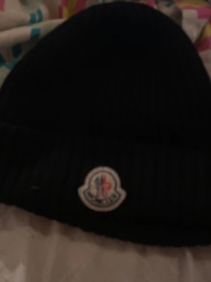 Svart ribbad mössa från Moncler - Säljer en svart ribbstickad mössa från Moncler med uppvikt kant och Monclers klassiska logga framtill. Mössan är tillverkad i mjukt material och har en stilren, enkel design som passar perfekt till kalla dagar.