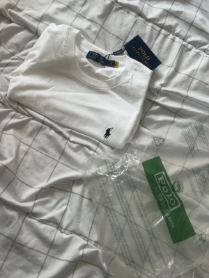 Vit t-shirt från Polo Ralph Lauren - Klassisk vit t-shirt från Polo Ralph Lauren med rund hals och ikoniska broderade loggan på bröstet. Tillverkad i mjuk bomull och har en normal passform. Perfekt basplagg för en clean och stilren look.
