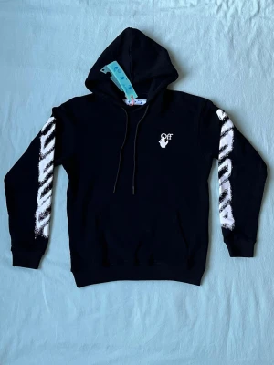 Svart Off-White hoodie med tryck - Svart hoodie från Off-White med vit logga på bröstet och ikoniska vita ränder med borstad effekt längs ärmarna. Klassisk huva med dragsko och känguruficka framtill. Perfekt för en streetwear-look.