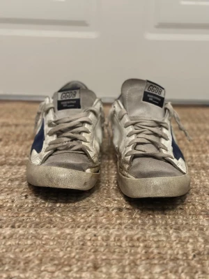 GGDB vita sneakers med blå stjärna - Säljer ett par GGDB sneakers i vitt läder med grå detaljer och en ikonisk blå stjärna på sidan. Skorna har klassisk låg profil, vita snören och GGDB-logga på plösen. Perfekta för dig som gillar streetstyle och vill ha en unik look.