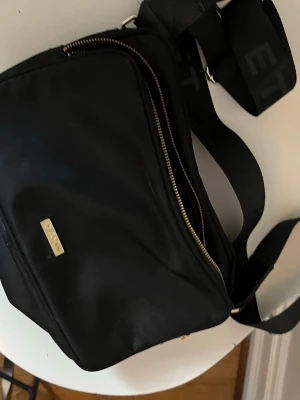 Svart axelväska från DAY ET - Snygg svart axelväska från DAY ET med justerbar bred axelrem där loggan är invävd. Väskan har guldfärgade dragkedjor och en metallbricka med DAY ET-logga framtill. Tillverkad i slitstarkt tygmaterial och har en modern, enkel design. Köpt för 950 men säljer för 300!