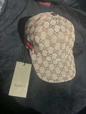 Gg keps - Snygg beige keps från Gucci med klassiskt GG-monogram över hela kepsen och gröna/röda ränder på sidan. Kepsen har böjd skärm och justerbar rem baktill. Perfekt accessoar för att lyfta din streetwear-look.