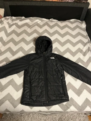 Svart vindjacka från The North Face - Snygg svart vindjacka från The North Face med huva och dragkedja framtill. Jackan har två sidofickor med dragkedja och diskret logga på bröstet. Tillverkad i lätt och slitstarkt material som står emot vind. Perfekt för blåsiga dagar och enkel att packa ner.