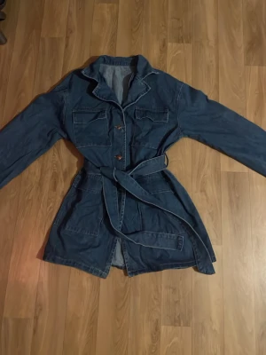 Blå jeansjacka  - Snygg blå jeansjacka med bälte i midjan och fyra stora fickor framtill. Jackan har klassisk krage, knappstängning och en avslappnad passform. Perfekt för dig som gillar en lite längre jeansjacka med coola detaljer.