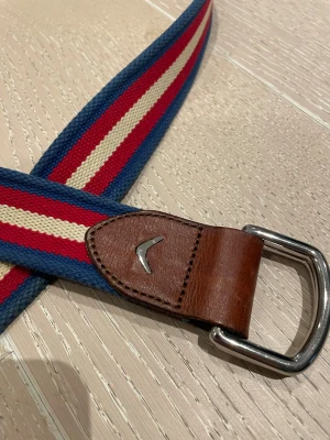 Randigt bälte med läderdetaljer från boomerange  - Snyggt randigt bälte i blått, rött och beige med bruna läderdetaljer och metallspänne. Perfekt accessoar för att lyfta din outfit med en sportig och retro känsla. Märke med metall-logga på lädret. Org pris: 700kr